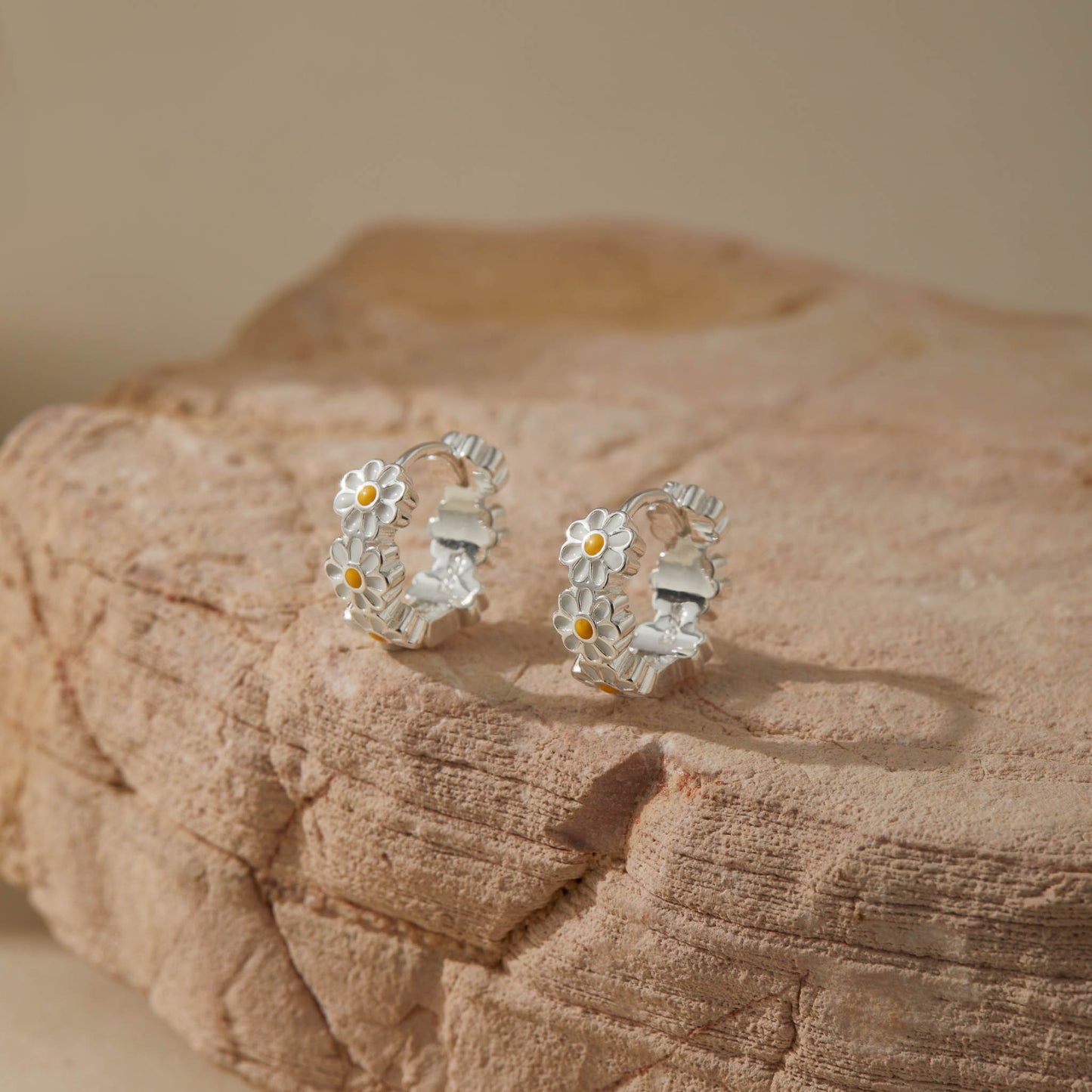 Pair of floral sterling silver daisy hoop earrings with enamel detail resting on beige rock.