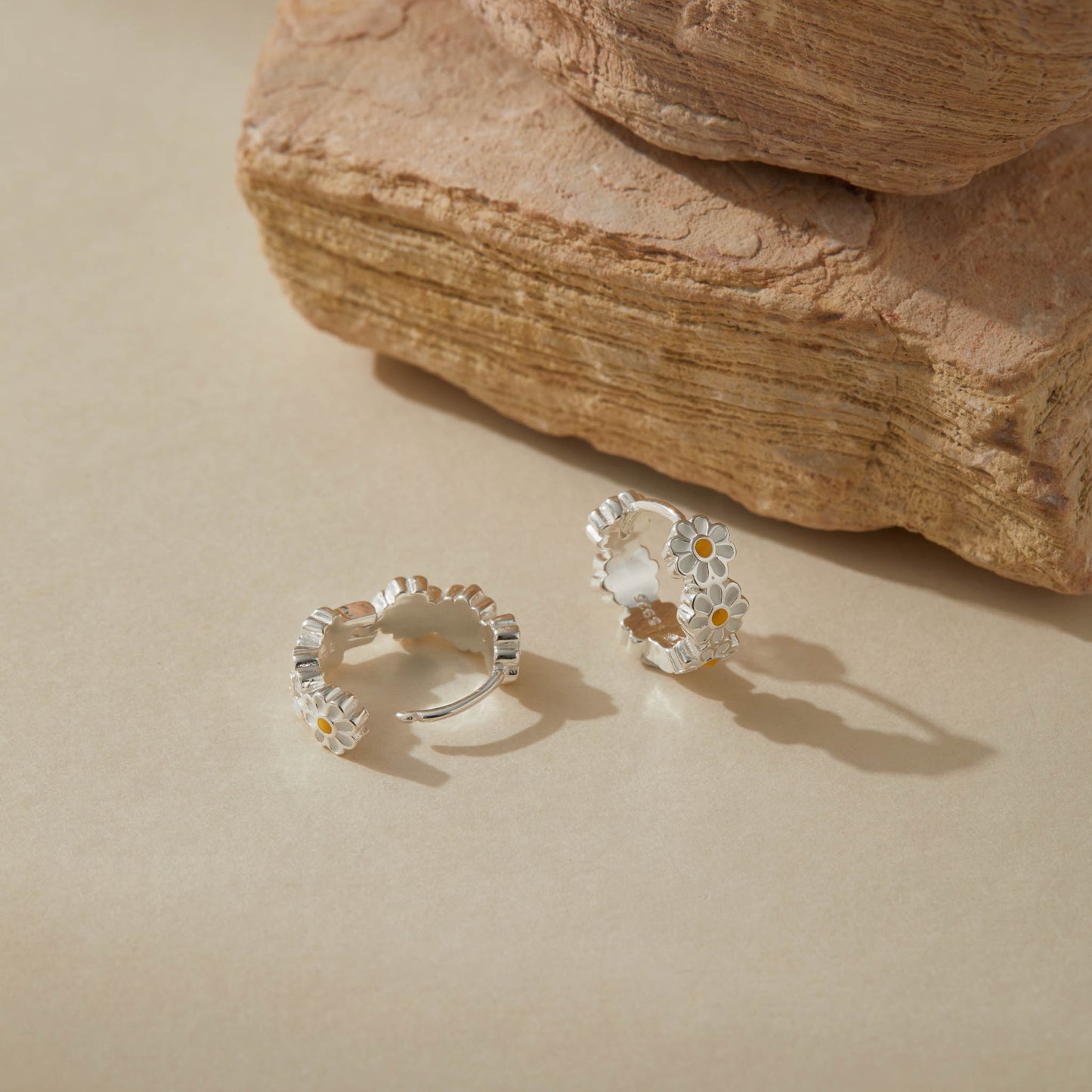 Close-up of sterling silver daisy hoop earrings with enamel flowers on neutral background.