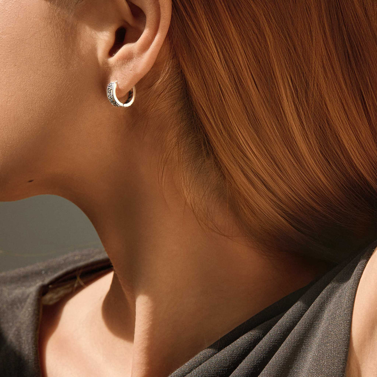 Side profile of a woman with red hair wearing a small sterling silver huggie hoop earring with an engraved gothic band