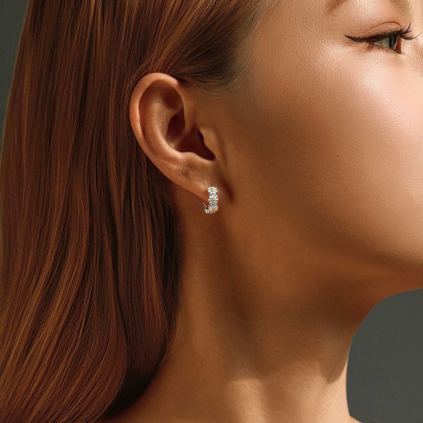 Close-up of woman wearing sterling silver daisy huggie earrings with white enamel petals and yellow centers.