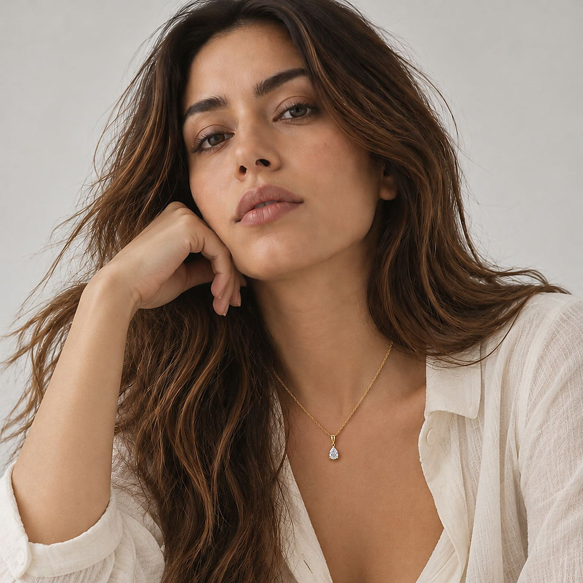 Woman resting her chin on her hand in an open cream gauze shirt, wearing a 14k gold plated pear pendant necklace