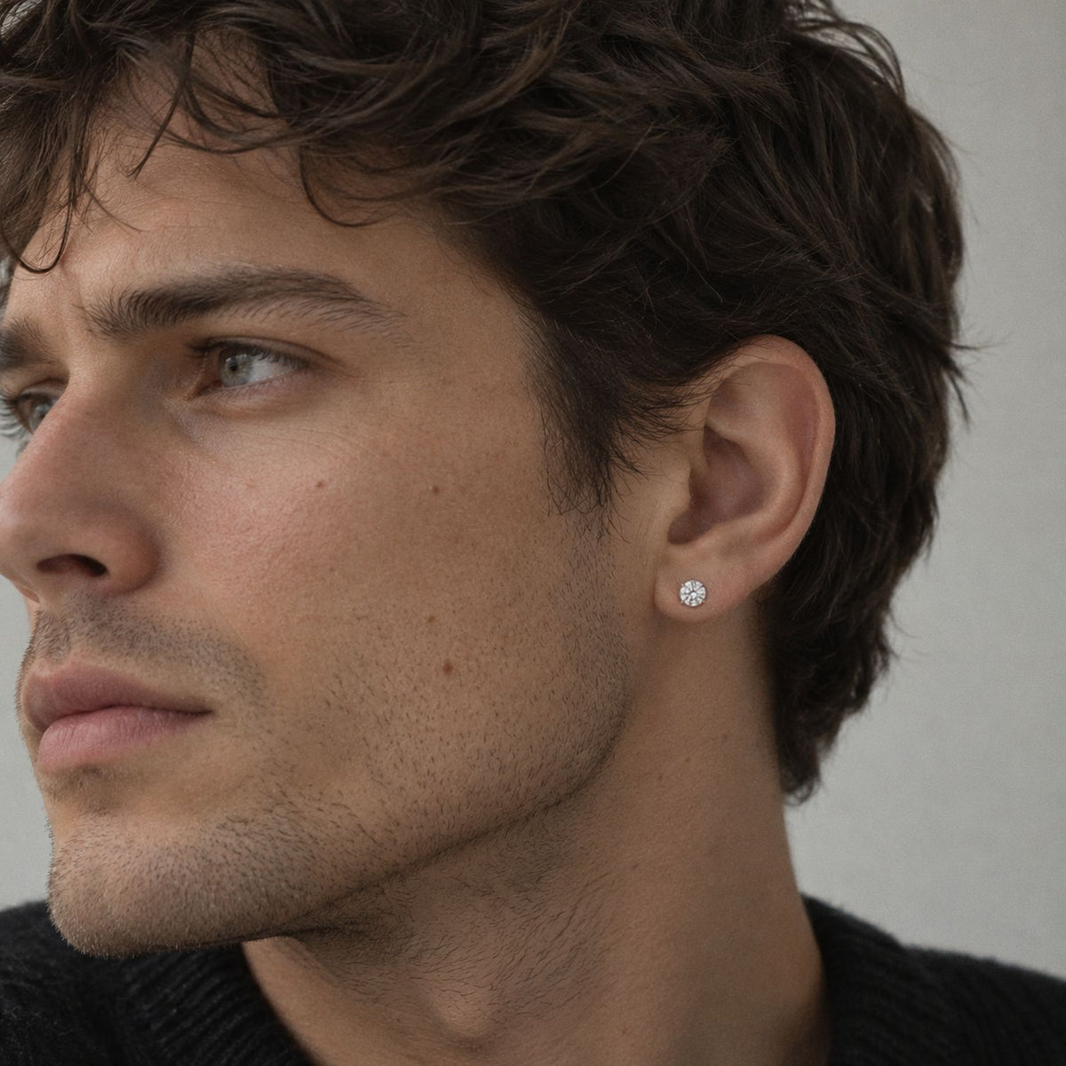 Dark haired man with stubble in a black knit, three quarter profile, wearing a round moissanite stud earring in silver