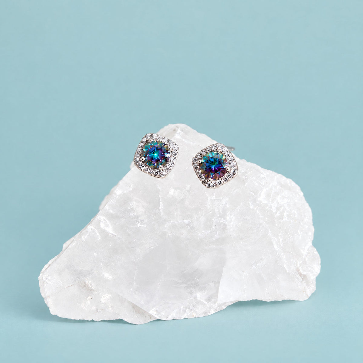 Rainbow blue moissanite halo stud earrings in sterling silver resting on a clear crystal against a soft teal backdrop