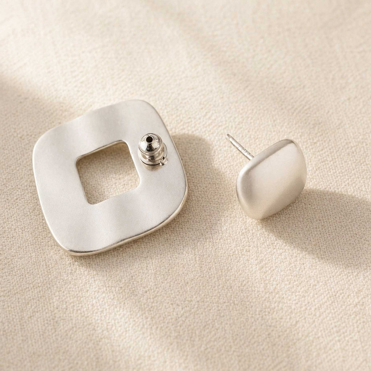 Matte sterling silver stud earrings shown from behind, open-square frame piece and cushion stud on cream linen
