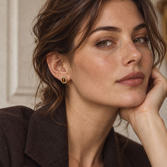 Woman with pinned-up brown hair wearing a gold-plated tiger eye stud earring, three-quarter view in a brown wool blazer