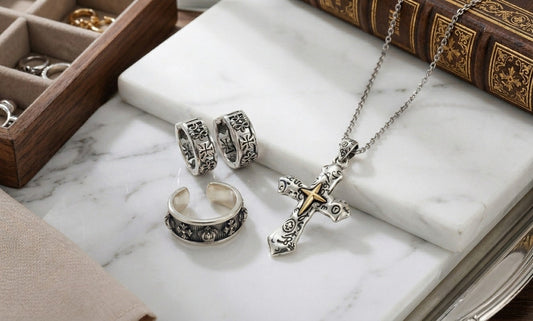 Oxidized gothic cross huggie earrings, antiqued sterling silver ring, and cross pendant necklace arranged on white marble surface with an old book