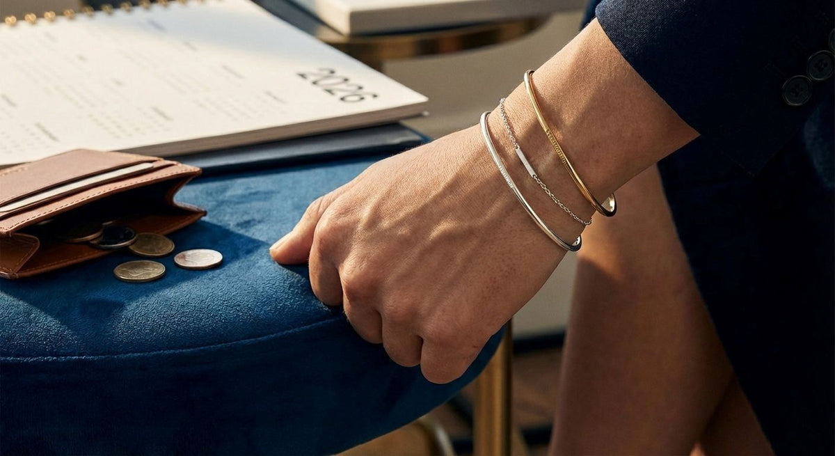A person wearing layered silver and gold bracelets next to a wallet, coins, and a 2026 calendar, illustrating jewelry investment trends.