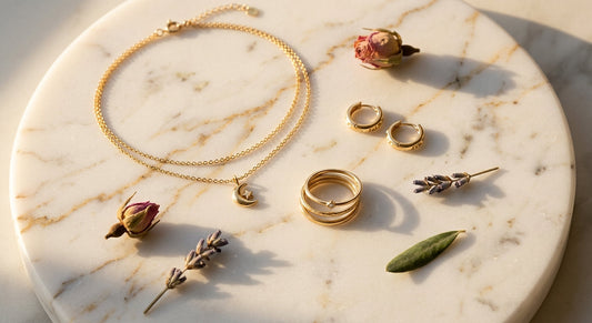 Flat lay of gold jewelry pieces including layered necklace, stacking rings and hoop earrings on marble surface with dried flowers, warm editorial lighting