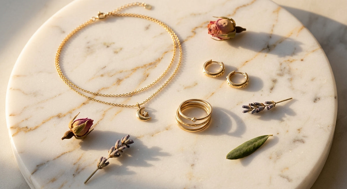 Flat lay of gold jewelry pieces including layered necklace, stacking rings and hoop earrings on marble surface with dried flowers, warm editorial lighting