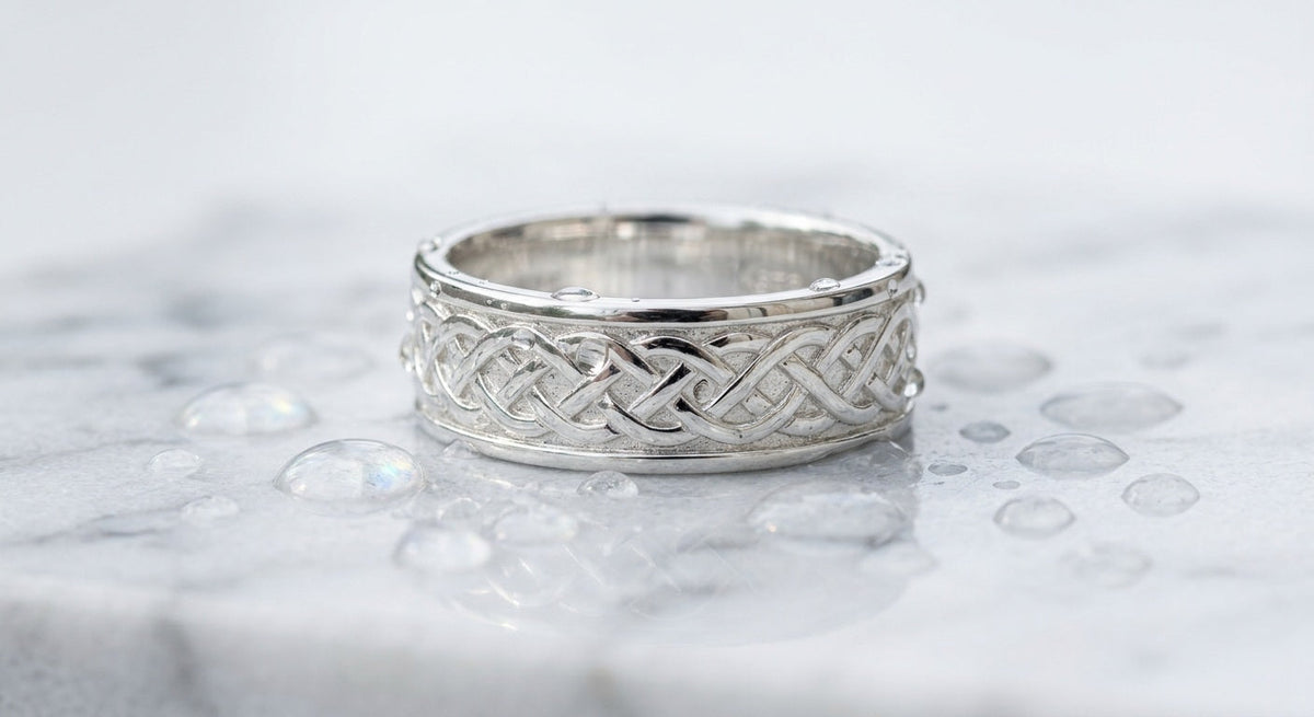 Polished sterling silver ring with intricate pattern on wet marble surface with water droplets, soft light
