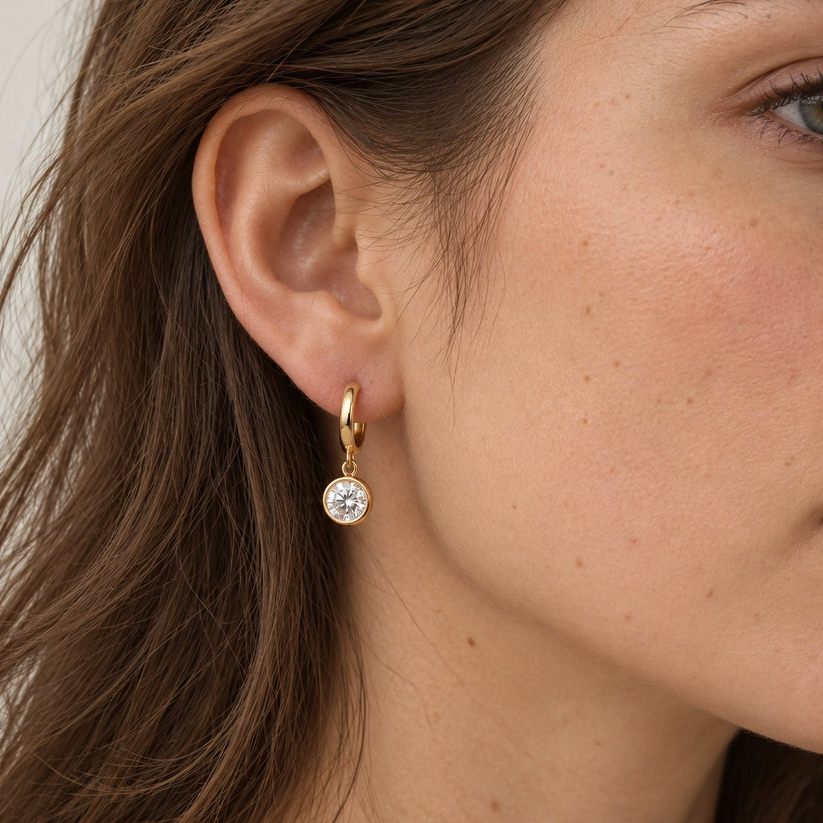 Woman with long brown hair in side profile wearing a small gold huggie earring with a round moissanite bezel drop