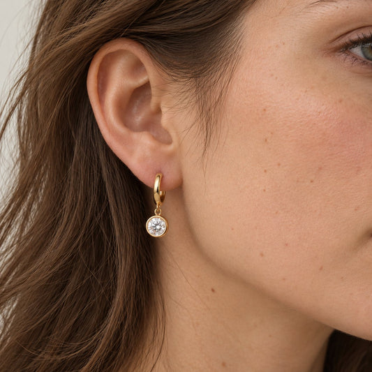 Woman with long brown hair in side profile wearing a small gold huggie earring with a round moissanite bezel drop