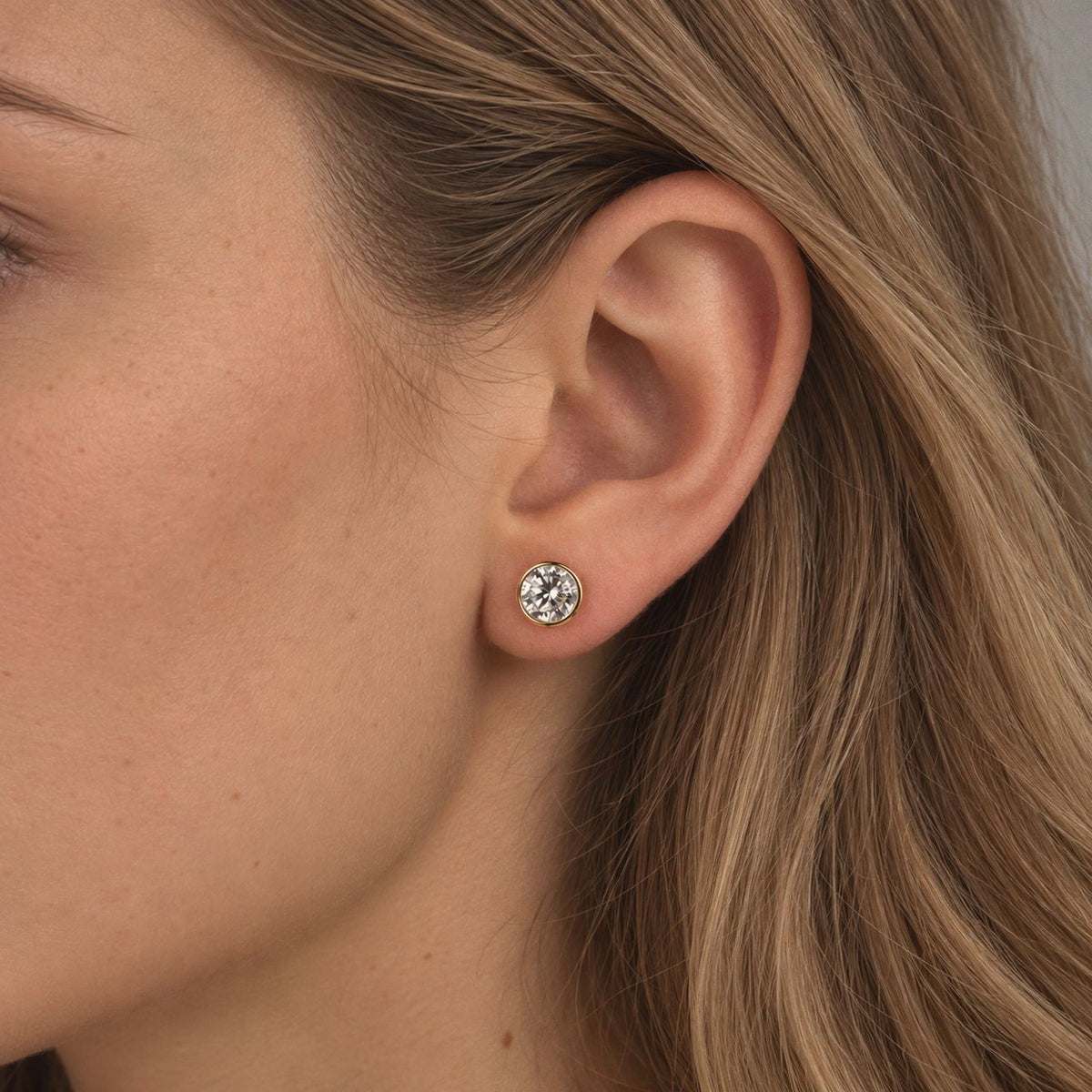 1 ct gold bezel moissanite stud worn on the lobe, side profile of a woman with light brown hair