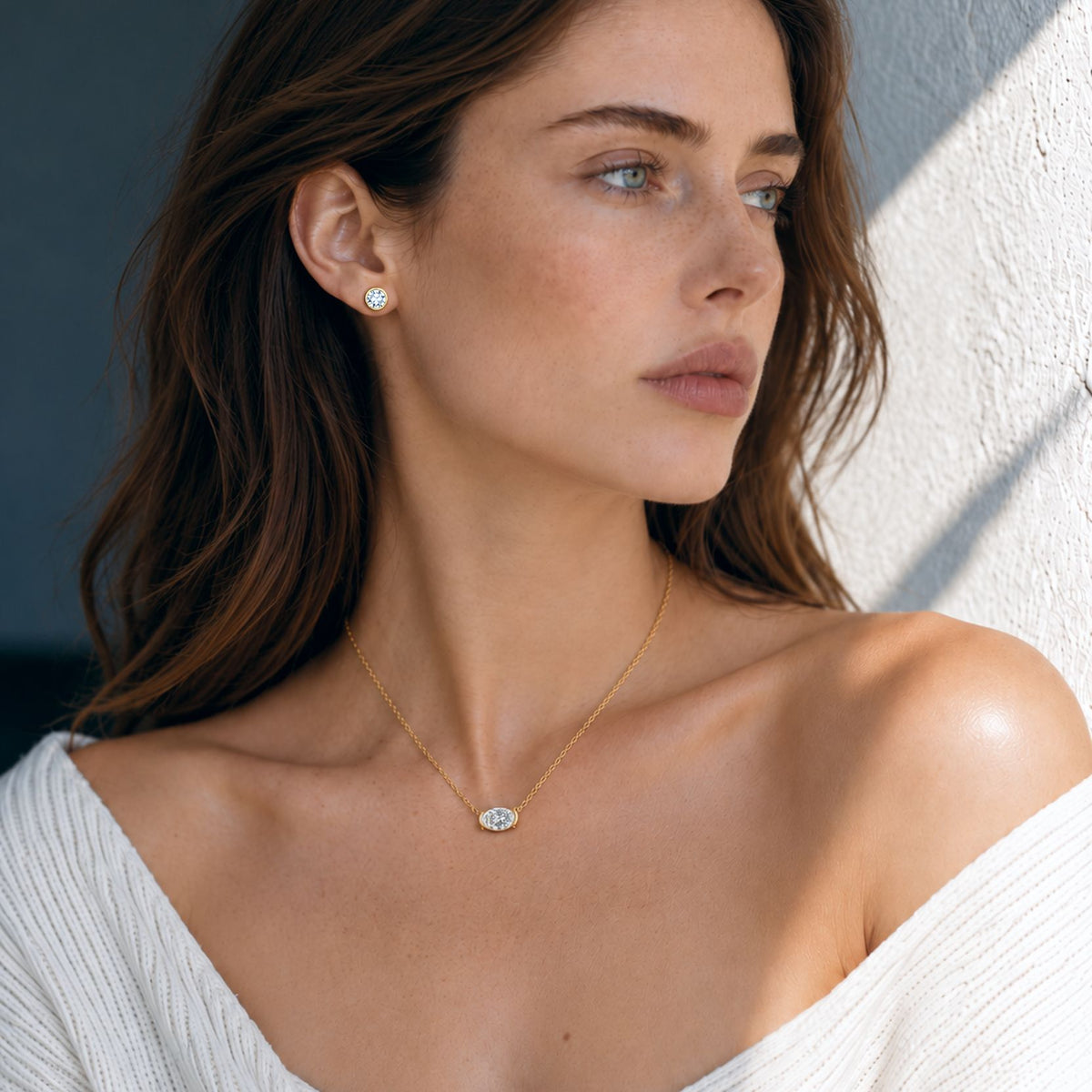 Woman wearing a 1 ct gold bezel moissanite stud earring with an oval pendant, looking toward a sunlit wall