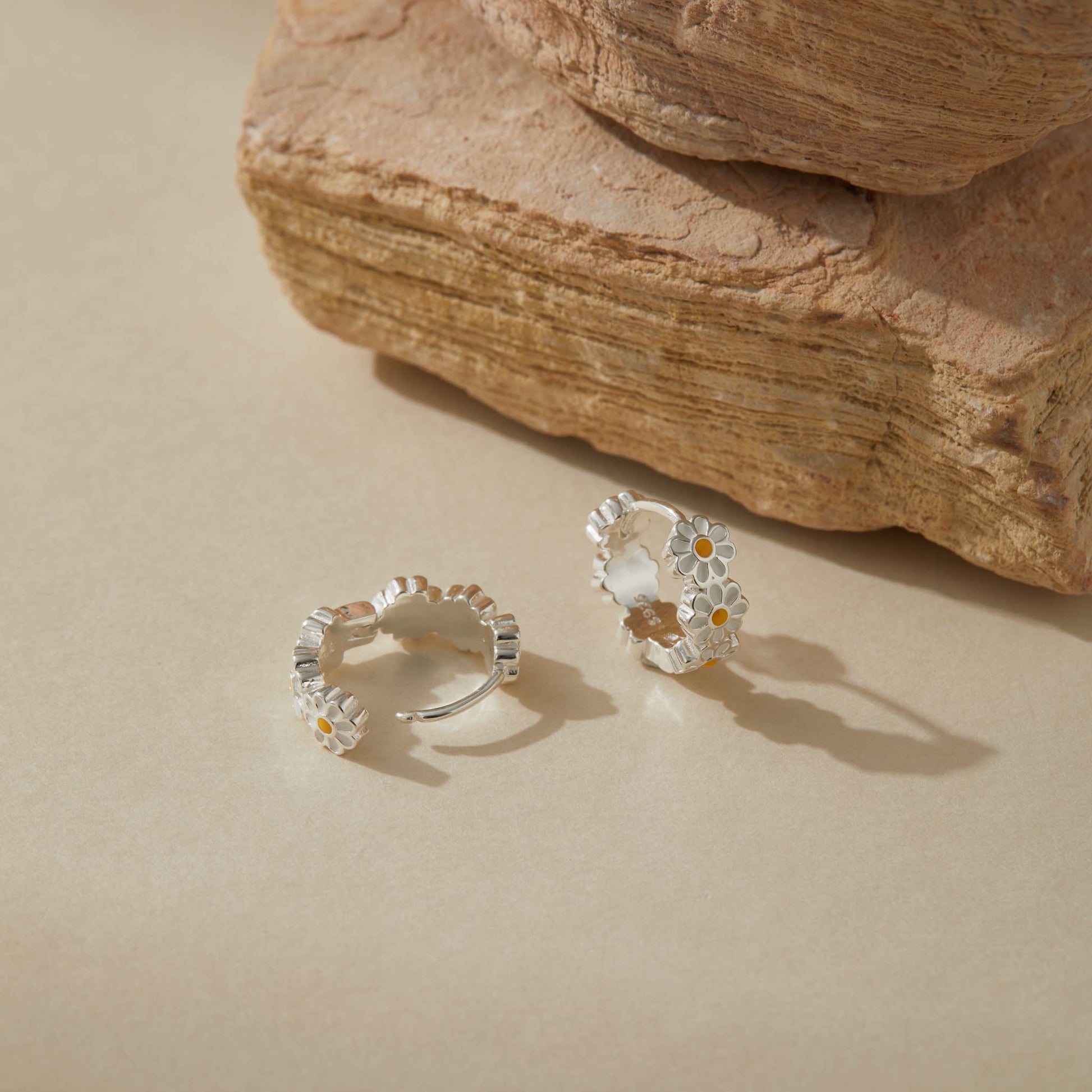 Close-up of sterling silver daisy hoop earrings with enamel flowers on neutral background.