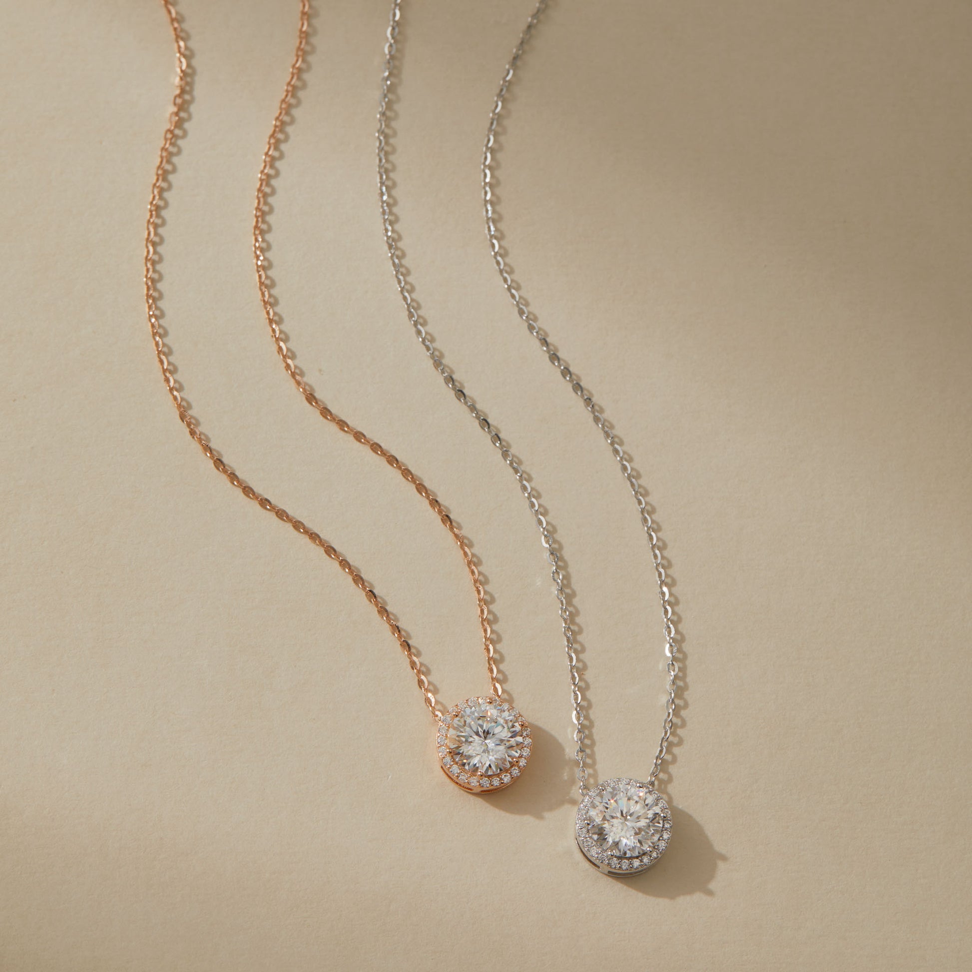 Rose gold and white gold moissanite halo pendant necklaces side by side on beige surface

