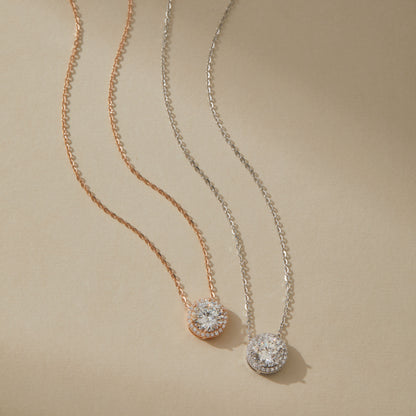 Rose gold and white gold moissanite halo pendant necklaces side by side on beige surface

