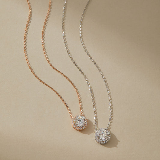 Rose gold and white gold moissanite halo pendant necklaces side by side on beige surface

