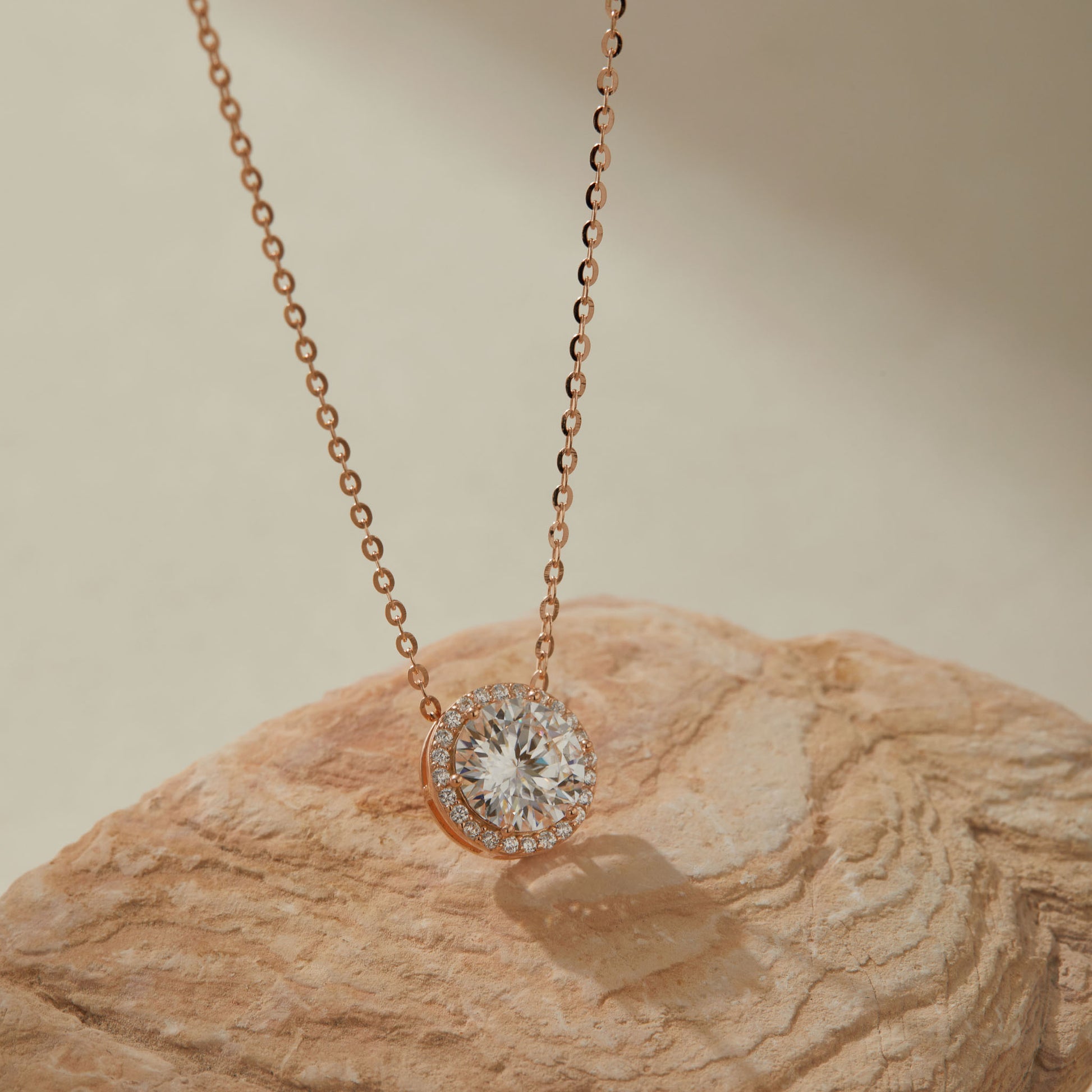 Rose gold-plated 2ct moissanite round halo pendant necklace on sandstone, close-up of pendant
