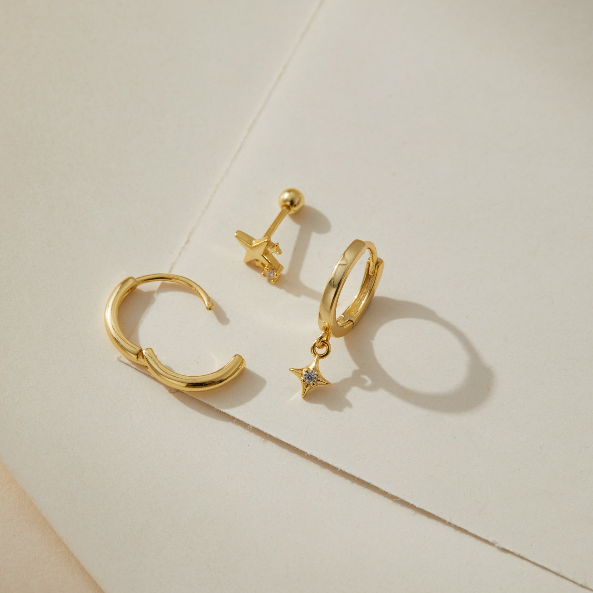 Gold plated star huggie earring set on cream paper, one hinged hoop open to show the closure