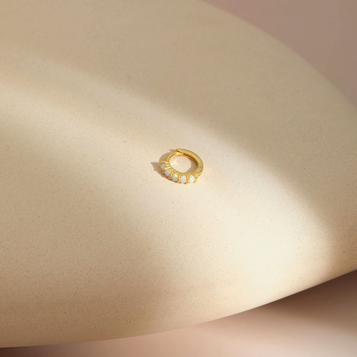 Single gold plated huggie hoop earring set with a row of white opals, lying on a sand-textured beige plinth