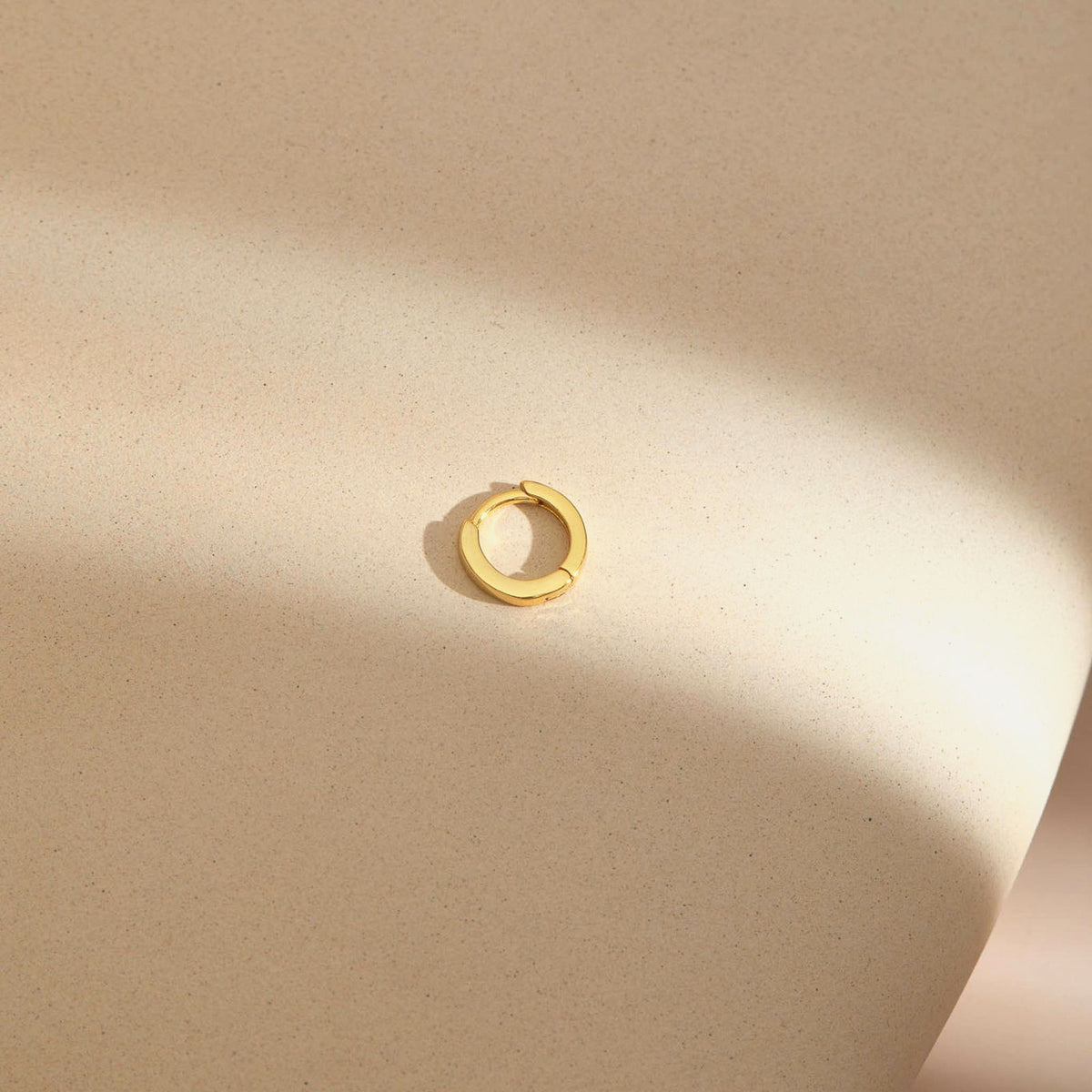 Plain gold plated huggie hoop earring from the opal set of three, on a sand-textured beige plinth in low sun