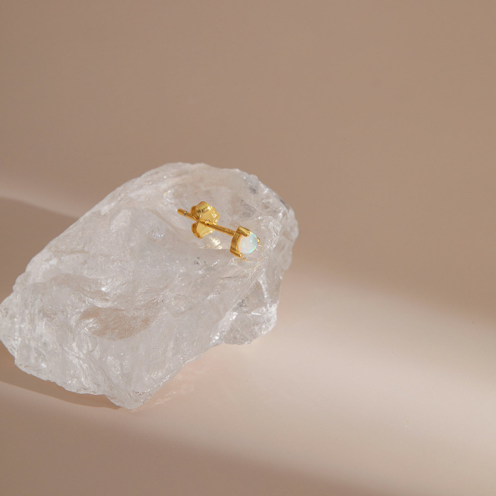 Gold plated opal stud earring with a single round opal in a claw setting, resting on raw quartz
