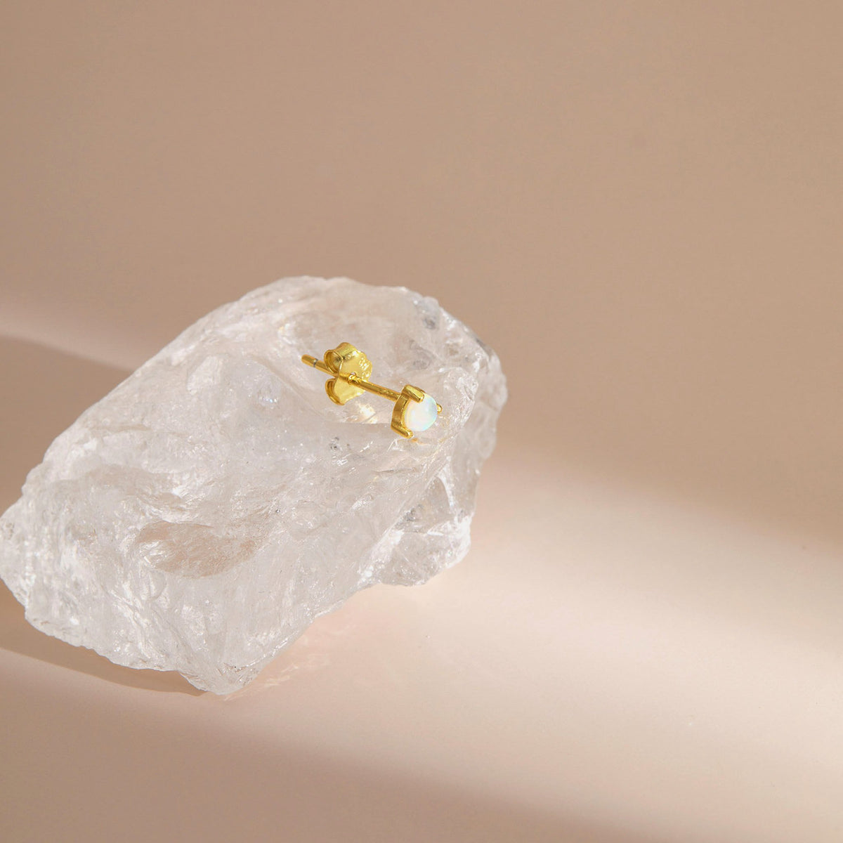 Gold plated opal stud earring with a round white opal, resting on a clear crystal against a soft blush-pink backdrop