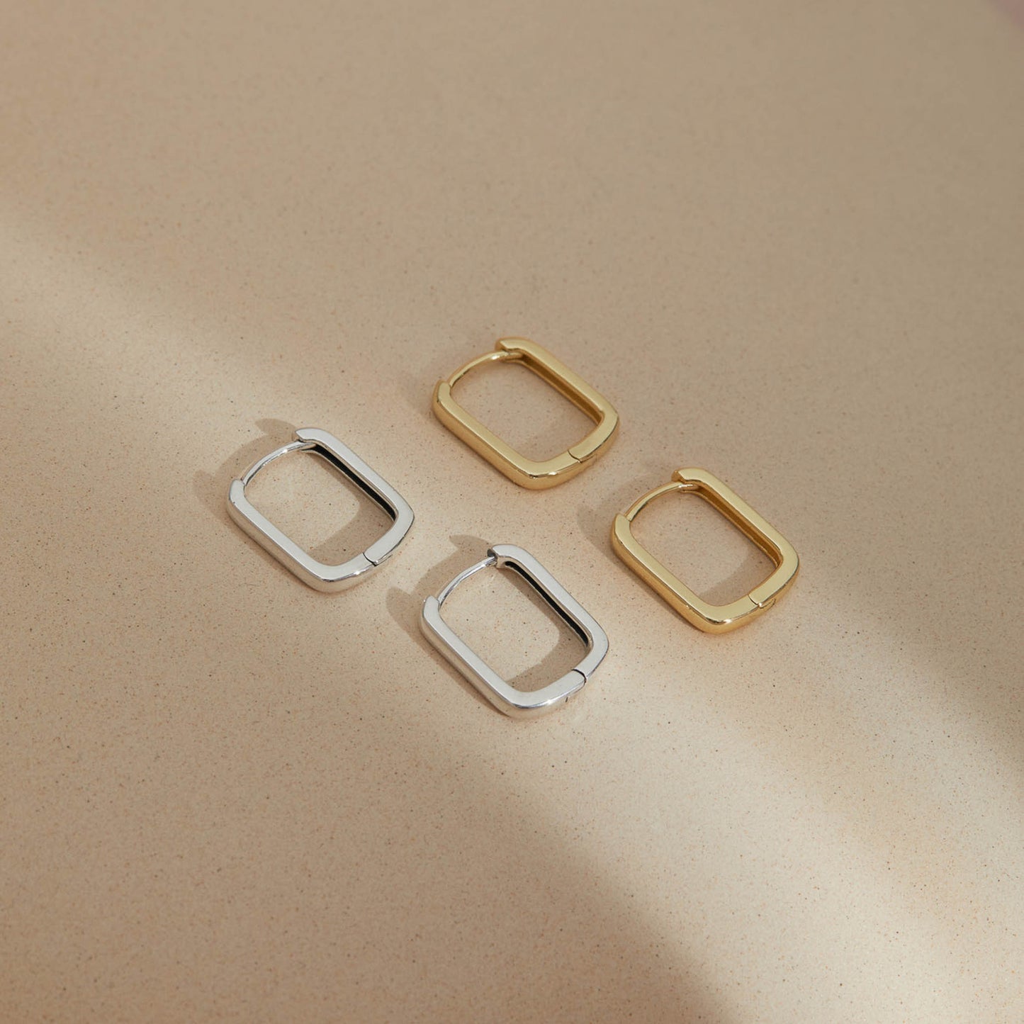 Minimalist rectangular huggie earrings in S925 sterling silver and gold plated displayed on a neutral surface, showcasing soft geometric design and refined craftsmanship.