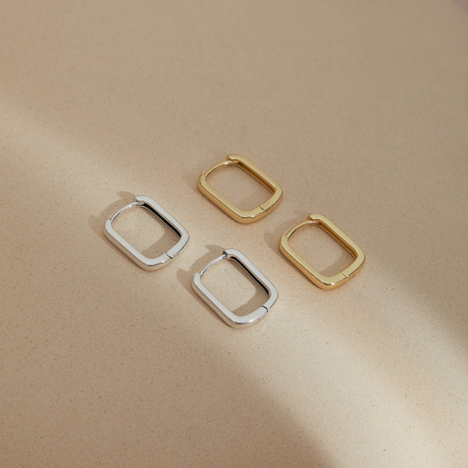 Minimalist rectangular huggie earrings in S925 sterling silver and gold plated displayed on a neutral surface, showcasing soft geometric design and refined craftsmanship.