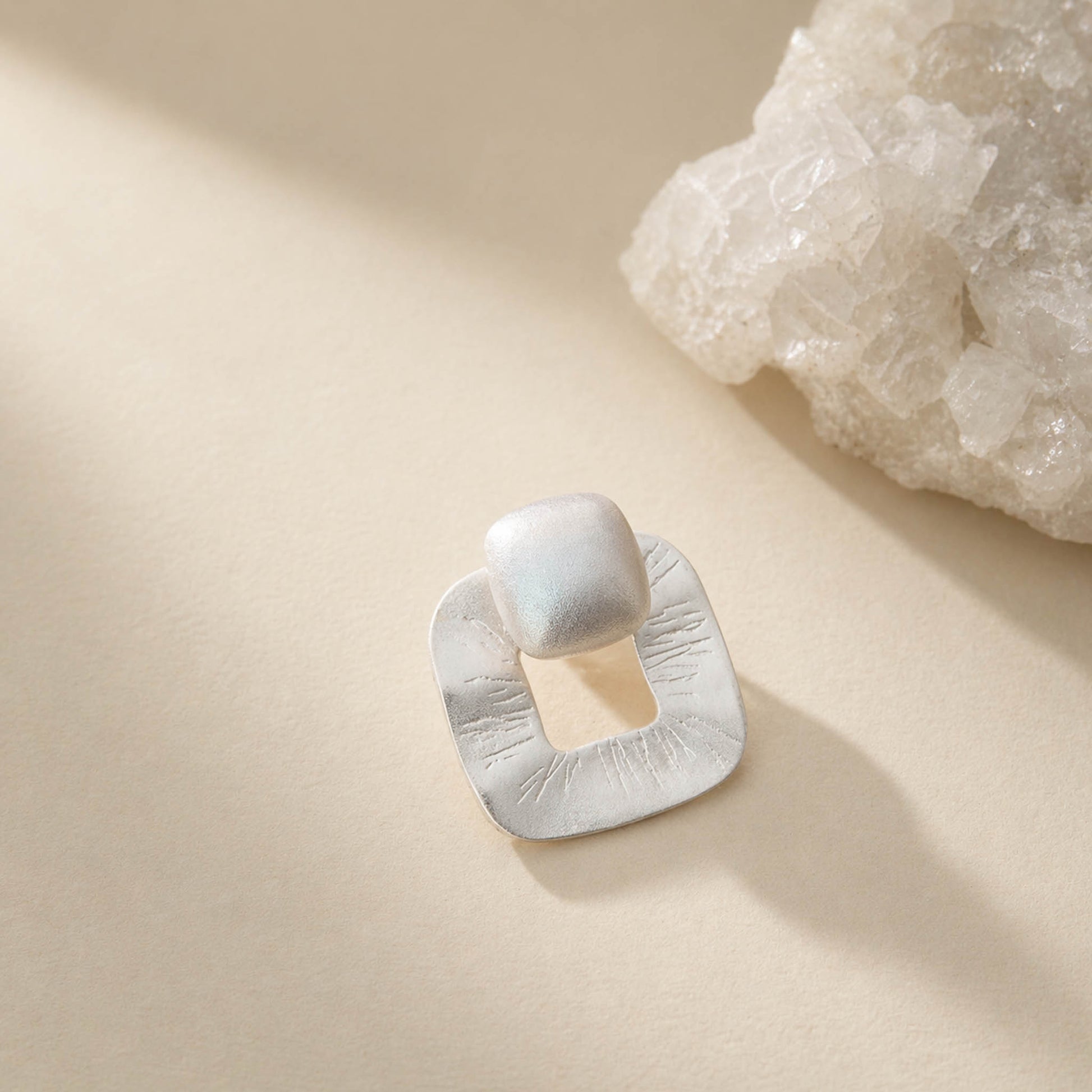 Brushed matte sterling silver stud earring with textured open-square frame and cushion top, on cream background