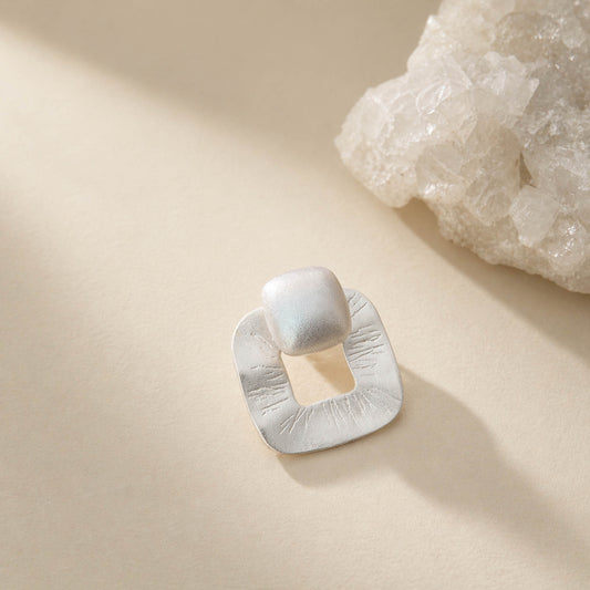 Brushed matte sterling silver stud earring with textured open-square frame and cushion top, on cream background
