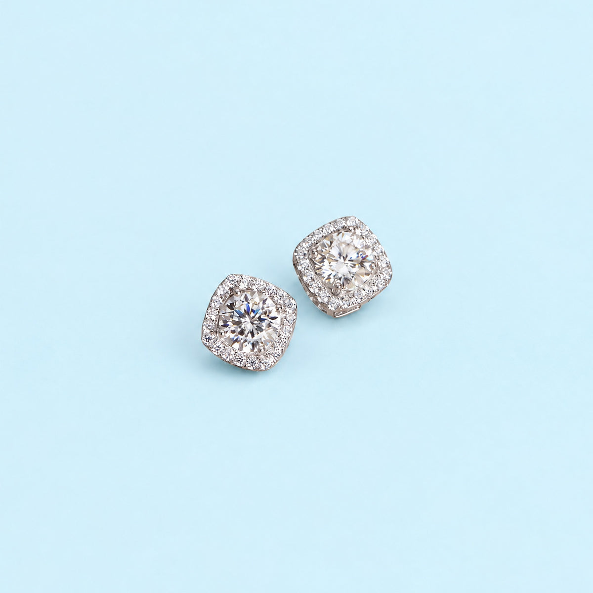 Colorless moissanite halo stud earrings in sterling silver lying flat on a pale ice blue backdrop, cushion halo