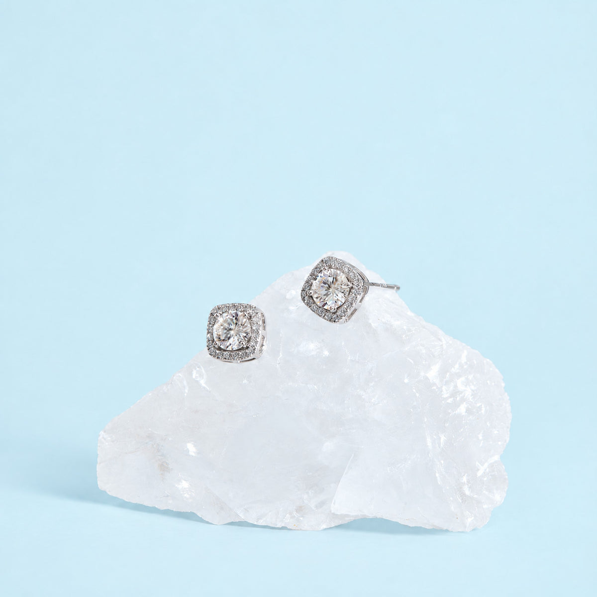 Colorless moissanite halo stud earrings in sterling silver resting on a clear crystal against a pale ice blue backdrop