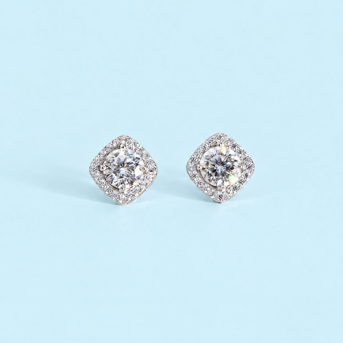 Pair of colorless moissanite halo studs facing the camera on a pale ice blue backdrop, round center stones