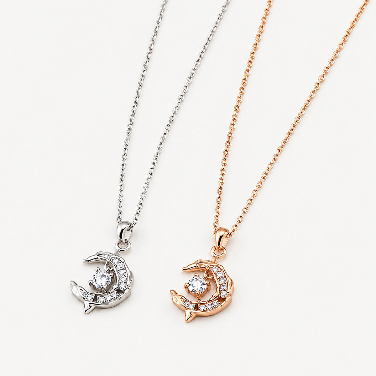 Two crescent moon dolphin necklaces, one 18k gold-plated and one sterling silver, pendants side by side on off-white