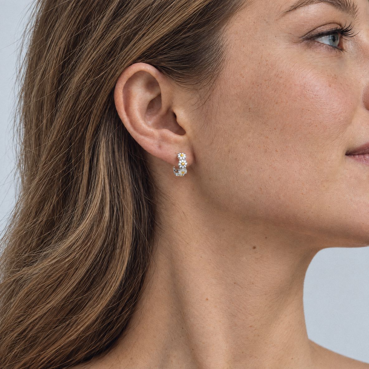 Woman with light brown hair wearing a sterling silver daisy huggie earring, side profile on a light grey backdrop