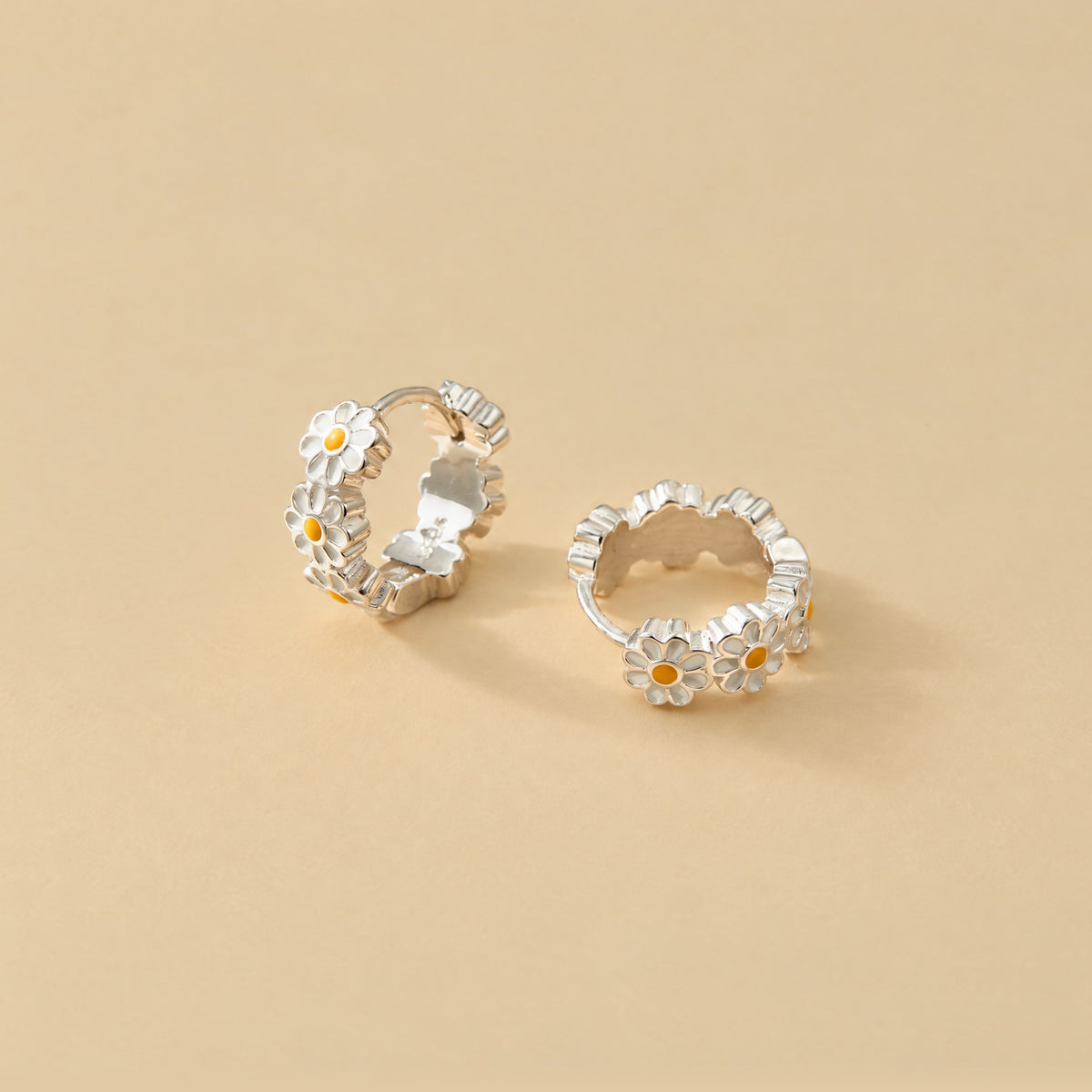 Sterling silver daisy huggie earrings on pale yellow, one lying flat to show the ring of daisies, one upright