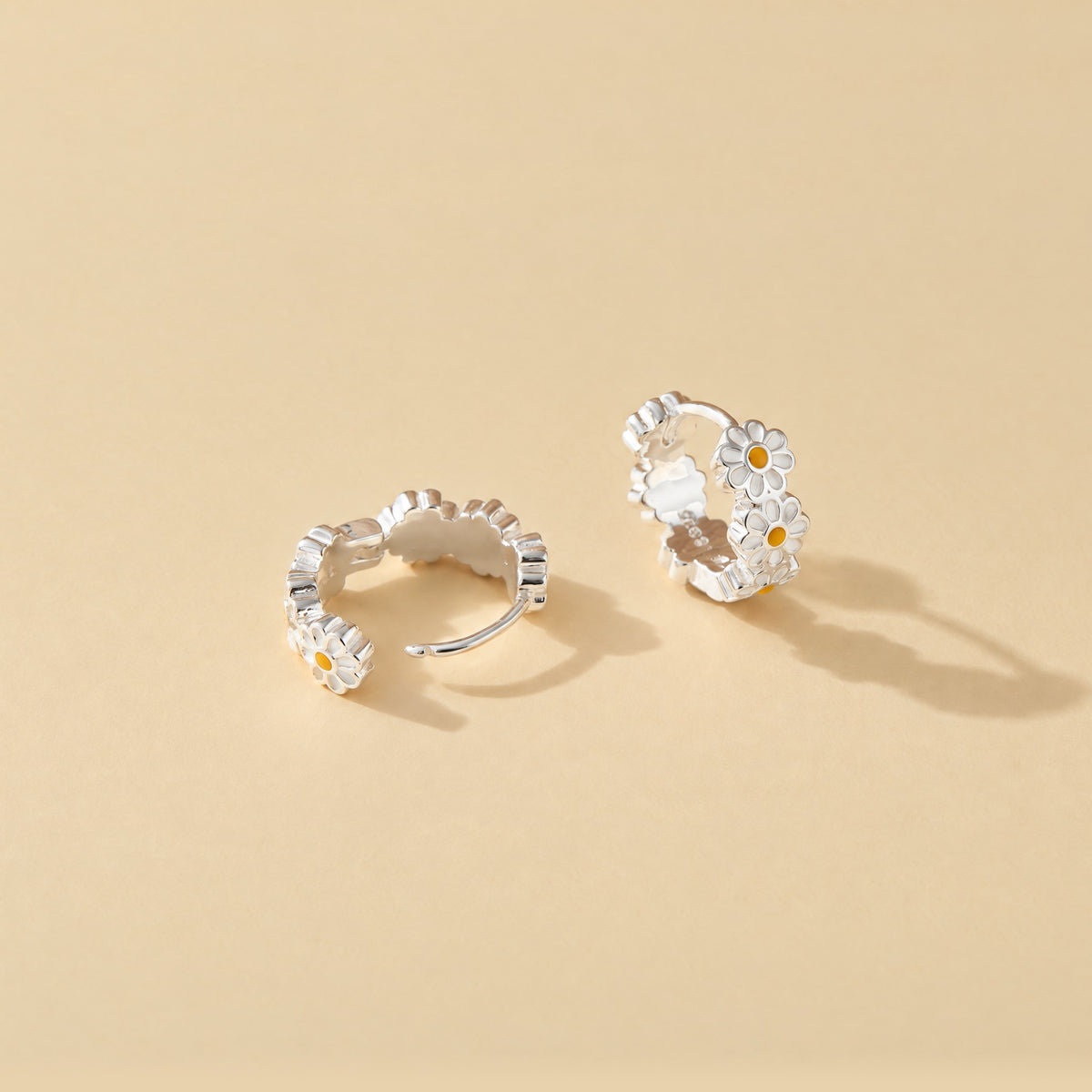 Sterling silver daisy huggie hoop earrings on pale yellow, one with the hinge open to show the post