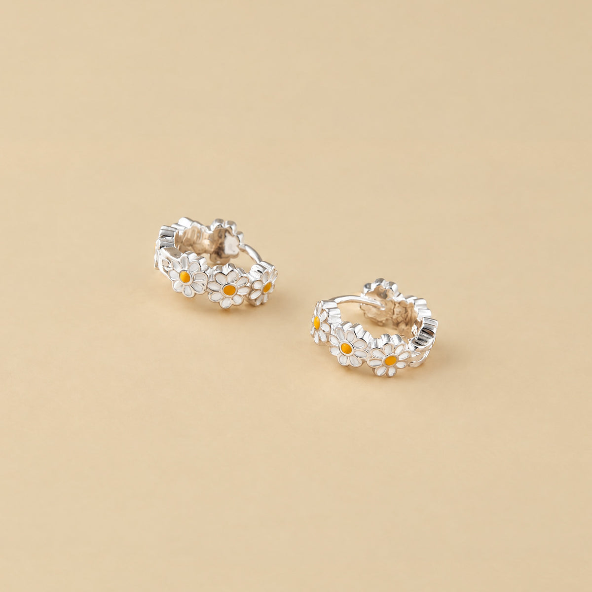 Sterling silver daisy huggie hoops lying side by side on a pale yellow backdrop, white petals and yellow centers