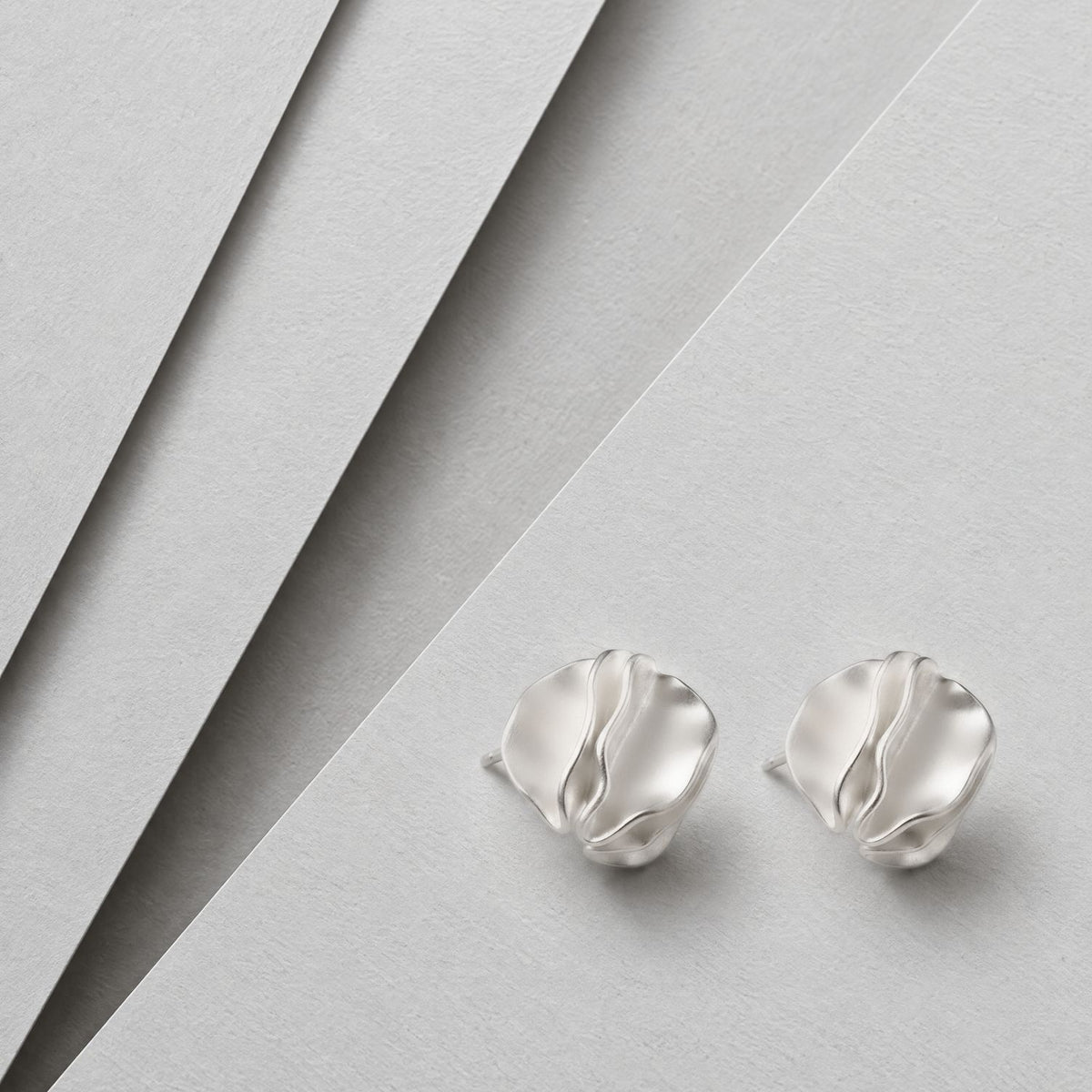 Pair of sterling silver folded petal stud earrings lying face up on layered grey card, brushed satin finish