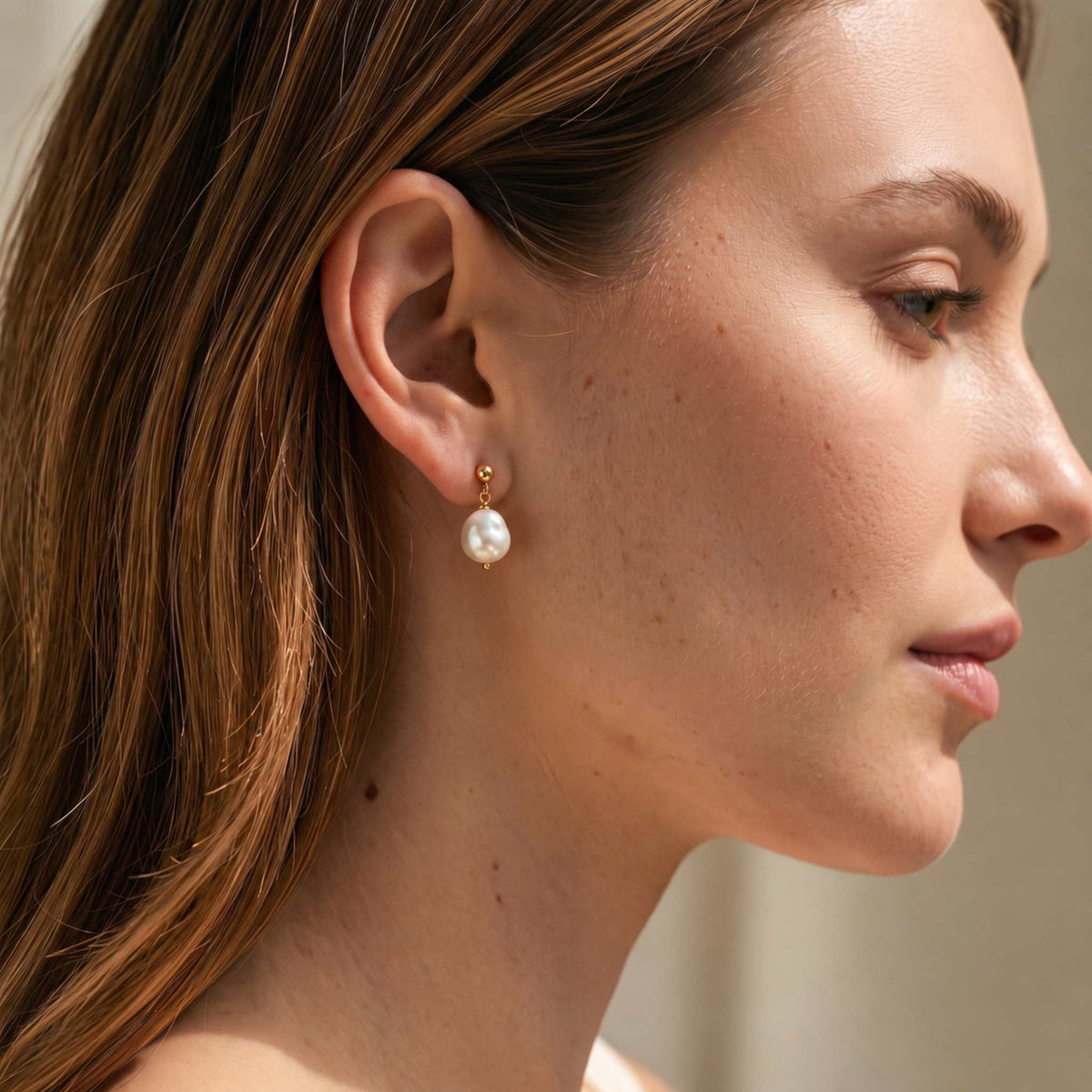 Woman in profile wearing a gold plated baroque freshwater pearl drop earring on her lobe, long brown hair tucked back
