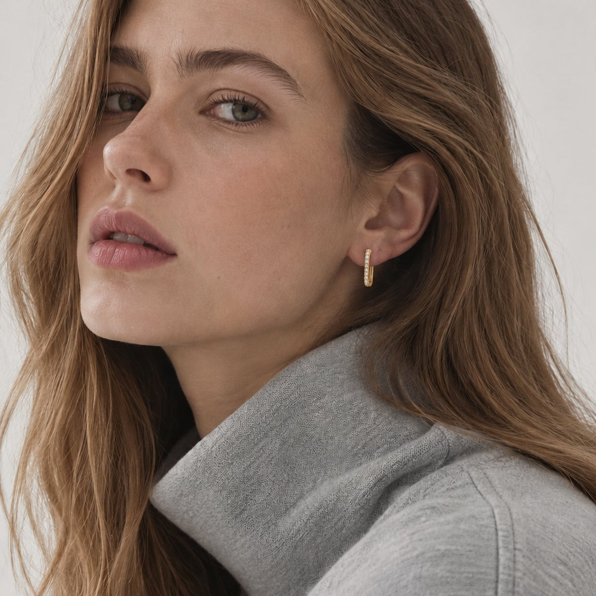 Portrait of a woman with long light brown hair in a grey sweater, wearing a gold plated U hoop huggie earring