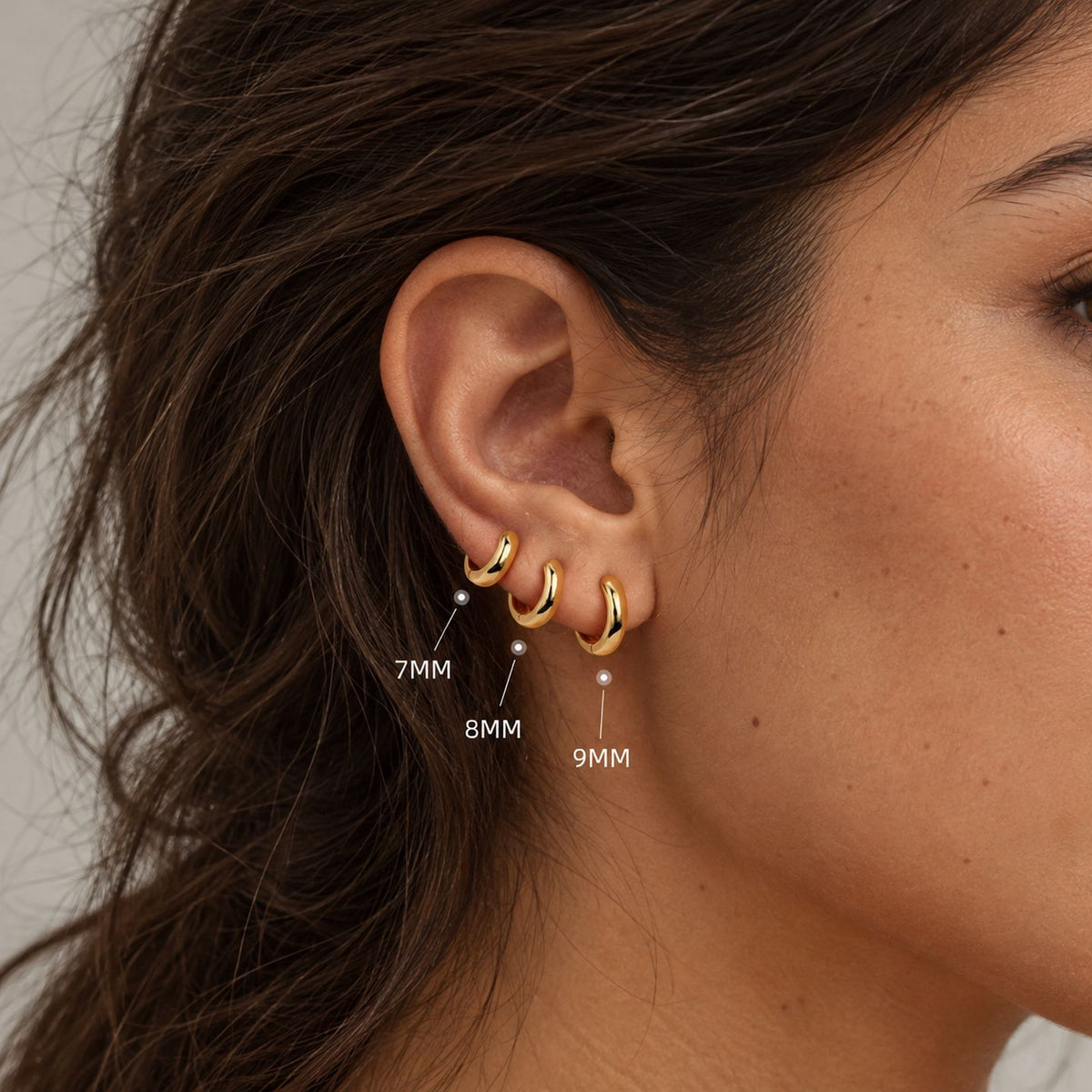 Woman with dark wavy hair wearing 7 mm, 8 mm and 9 mm gold dome huggies in a row on her earlobe, sizes labeled