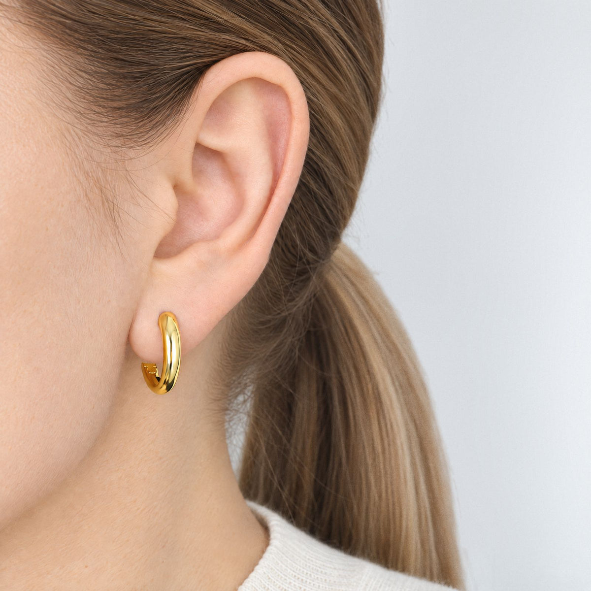 Woman with a blonde ponytail wearing a gold-plated tapered huggie hoop on her earlobe, side view in an ivory knit