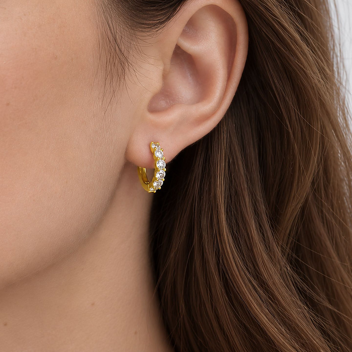 Close crop of a woman's ear and cheek, long brown hair, with a 14k gold plated moissanite huggie hoop worn in the lobe