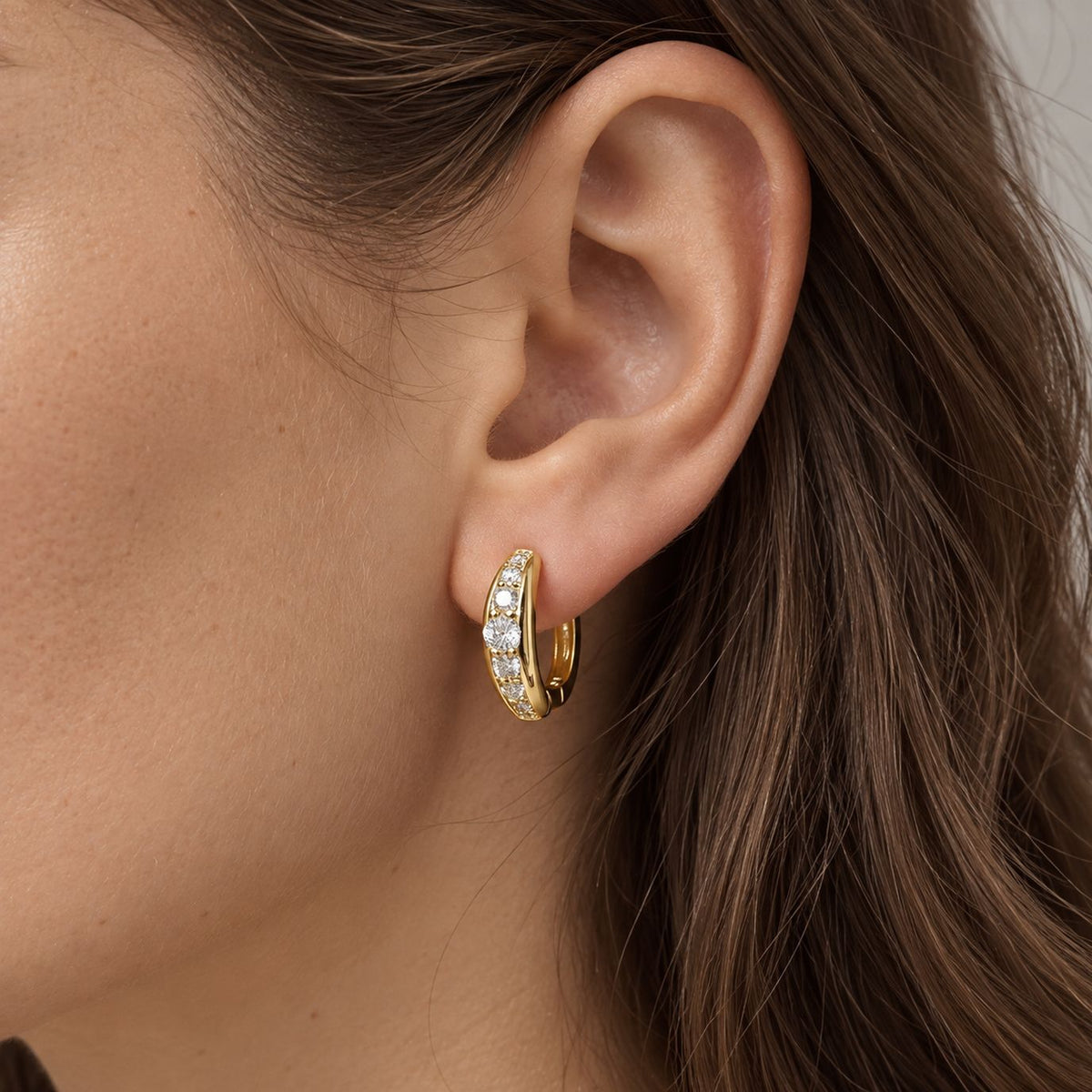 Gold moissanite huggie hoop earring worn on the earlobe, close side view of a woman with long brown hair