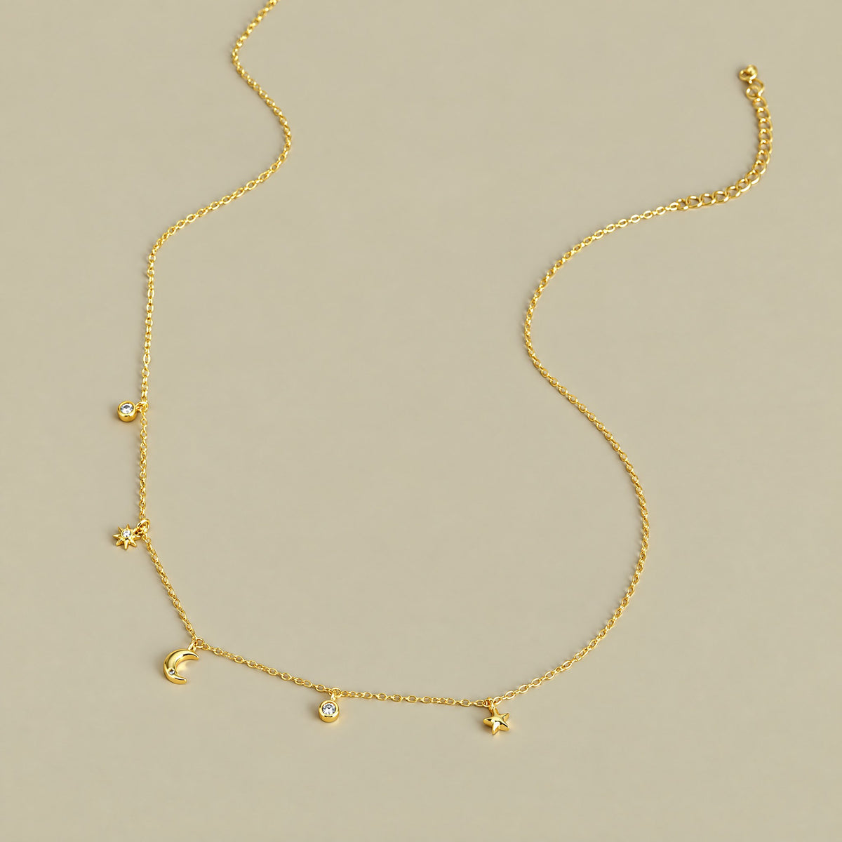 Gold plated moon and star charm necklace laid out in full on a pale khaki backdrop, charms spaced along the chain