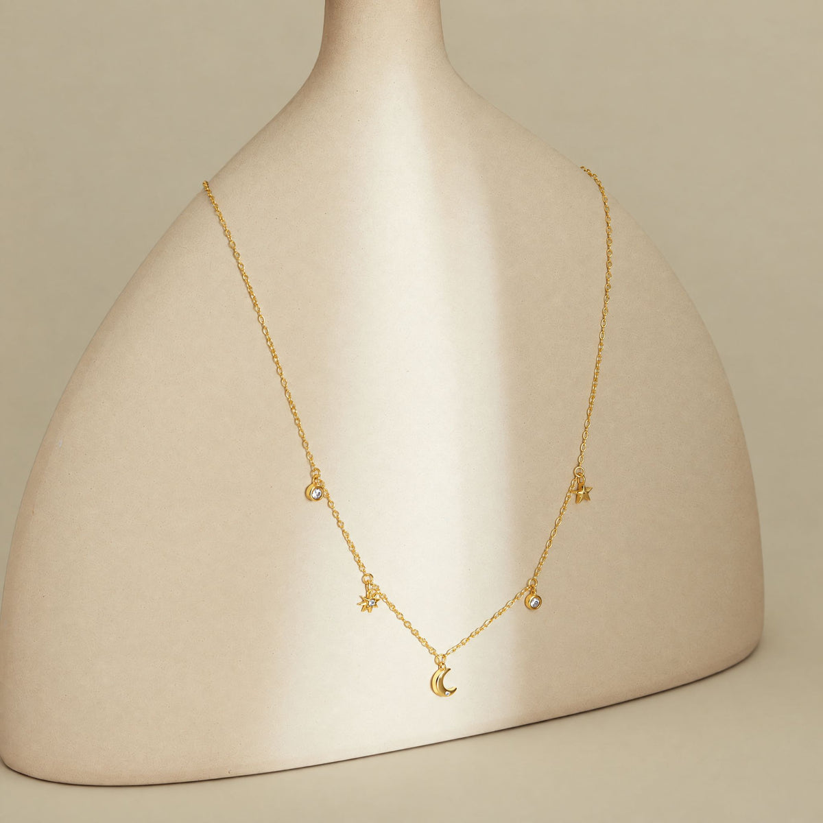 Gold plated moon and star charm necklace hanging on a cream sculptural stand against a pale khaki backdrop