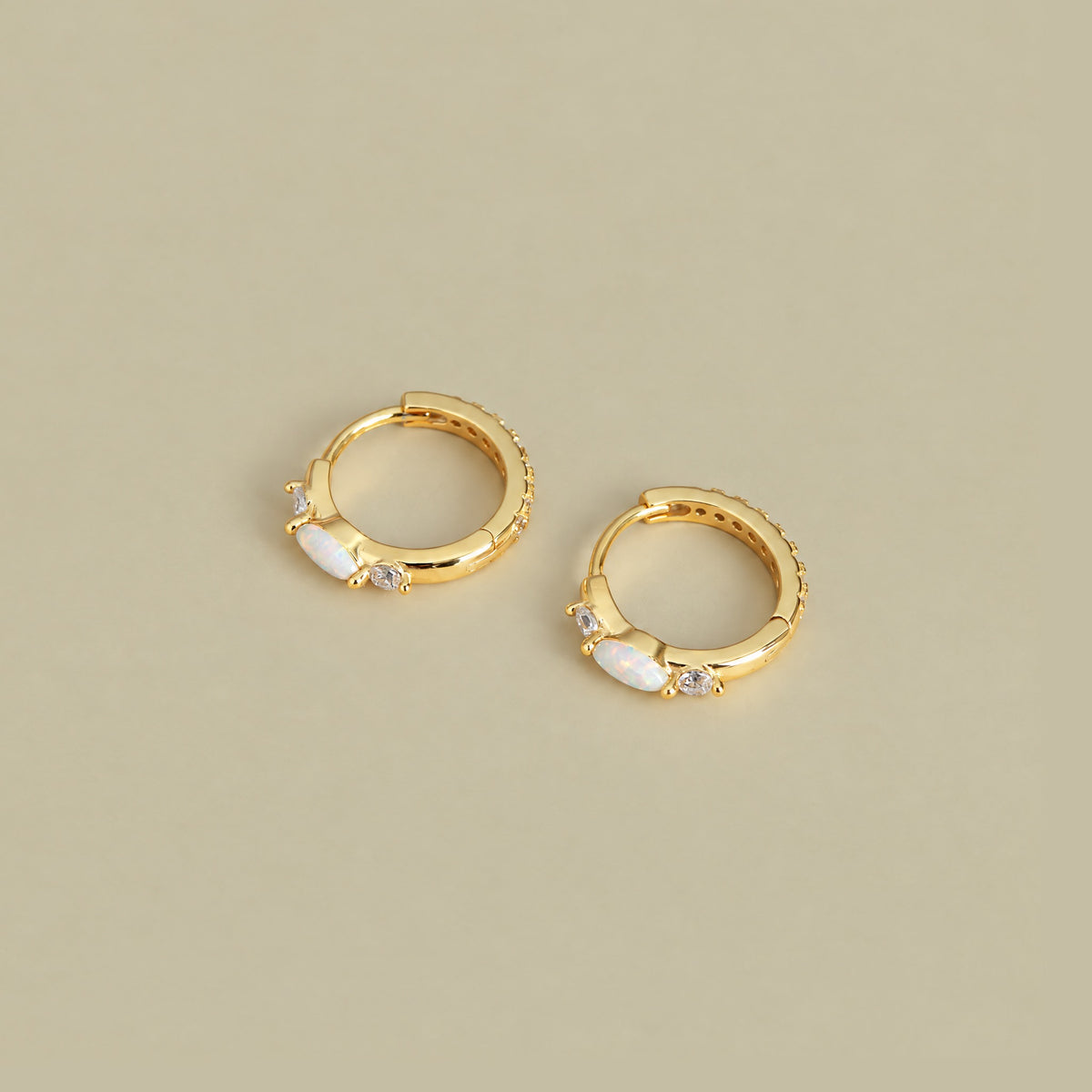Pair of gold plated opal huggie hoop earrings lying flat on a pale khaki backdrop, marquise opal and cubic zirconia